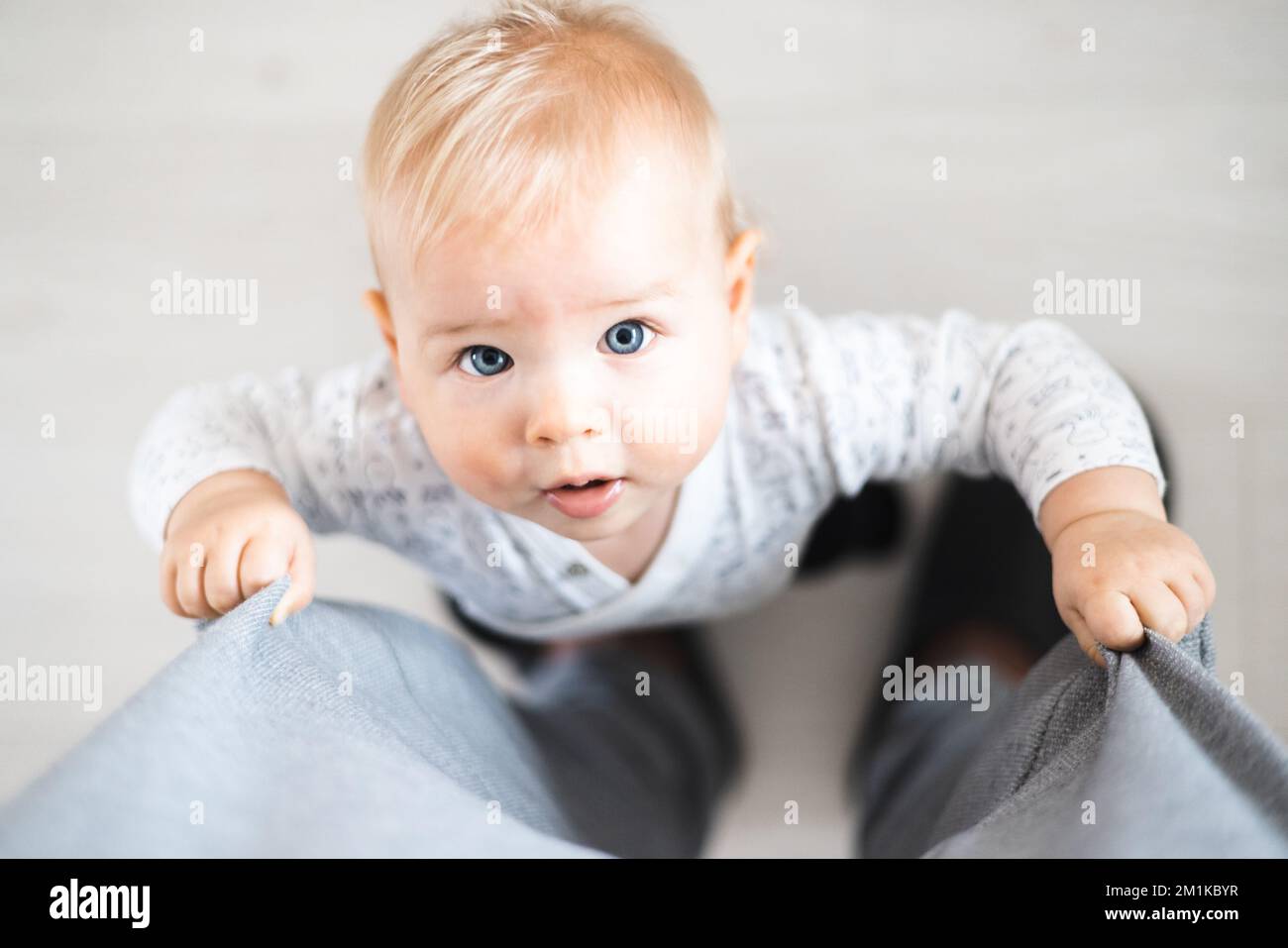Blick von oben auf den fröhlichen kleinen Jungen, der die ersten Schritte unternimmt und zu Hause an Vaters Trainingshose festhält. Süßer kleiner Junge, der Laufen lernt. Stockfoto