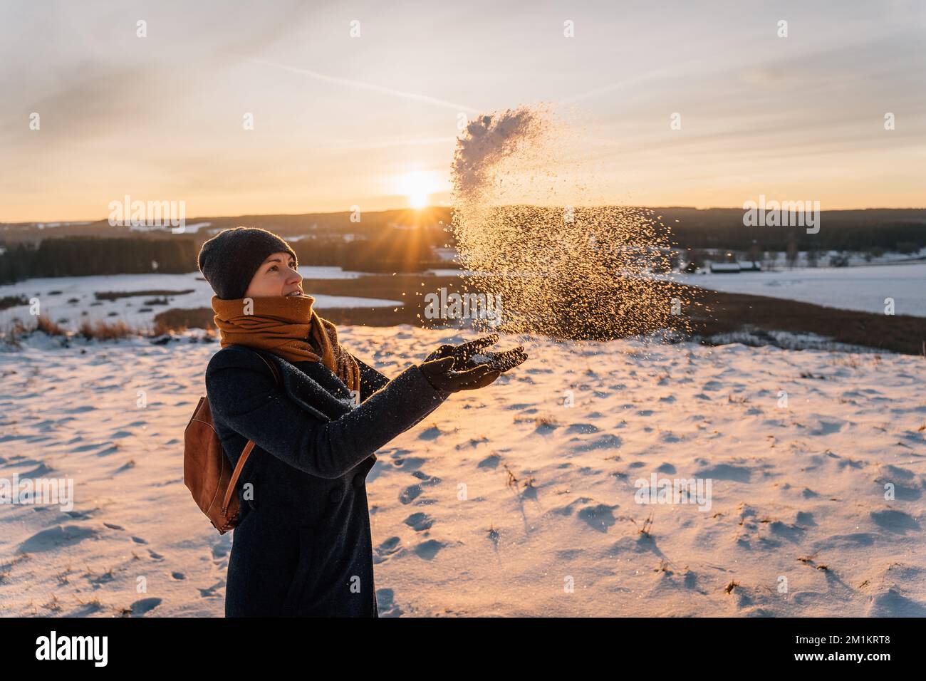 Eine Frau auf einem schneebedeckten Feld wirft Schnee mit ihren Händen vor dem Hintergrund eines Sonnenuntergangs Stockfoto