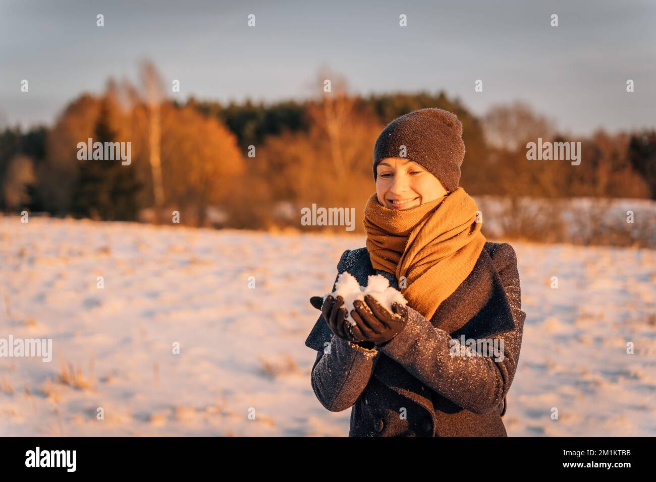 Eine lächelnde Frau mit Schal und Hut hält im Winter eine Handvoll Schnee Stockfoto