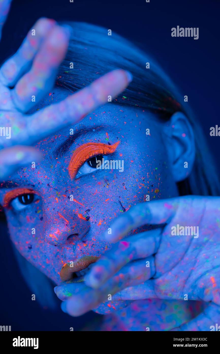 Frau in Neon-Make-up und helle Farbspritzer posieren mit verschwommenen Händen in der Nähe des Gesichts isoliert auf dunkelblauem Stockbild Stockfoto