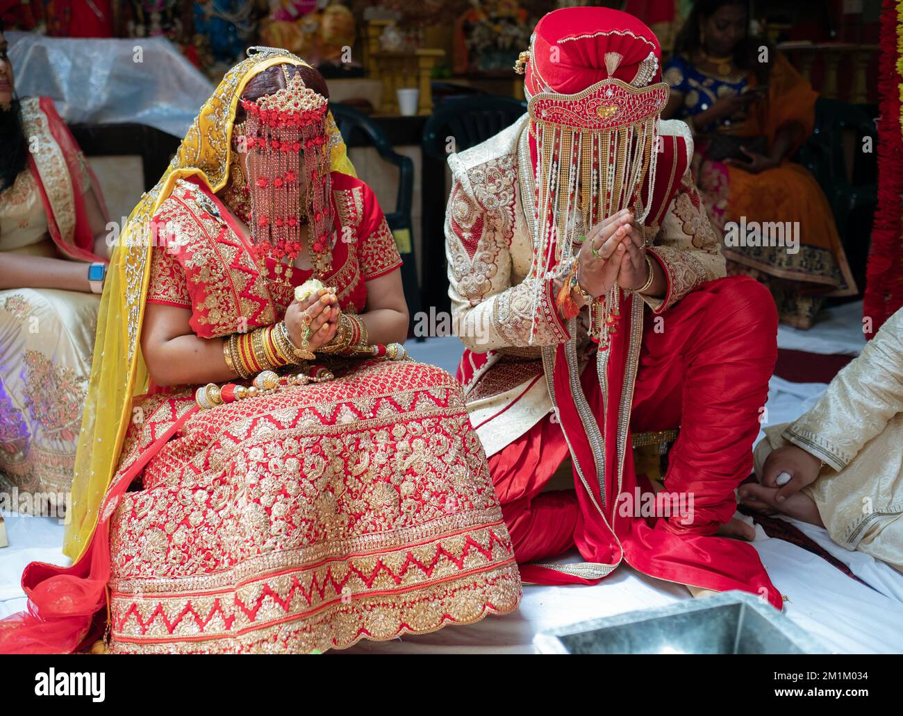 Bei einer Hindu-Hochzeit sitzen Braut und Bräutigam mit gefalteten Händen und tragen Sehra-Kopfkissen. In Queens, New York City. Stockfoto