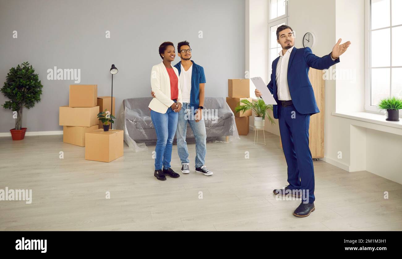 Professioneller männlicher Immobilienmakler zeigt jungen afroamerikanischen Paar eine helle neue Wohnung. Stockfoto