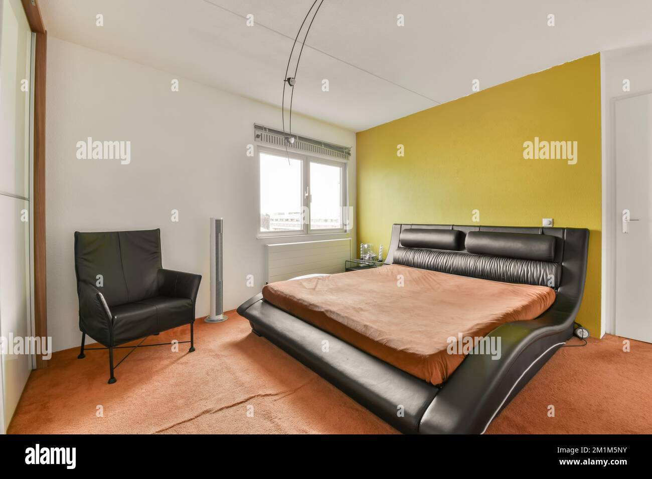 Ein Schlafzimmer mit einem orangefarbenen Bett und schwarzem Ledersessel im Zimmer ist hellgelbe Wände, weiße Verzierungen um das Fenster herum Stockfoto