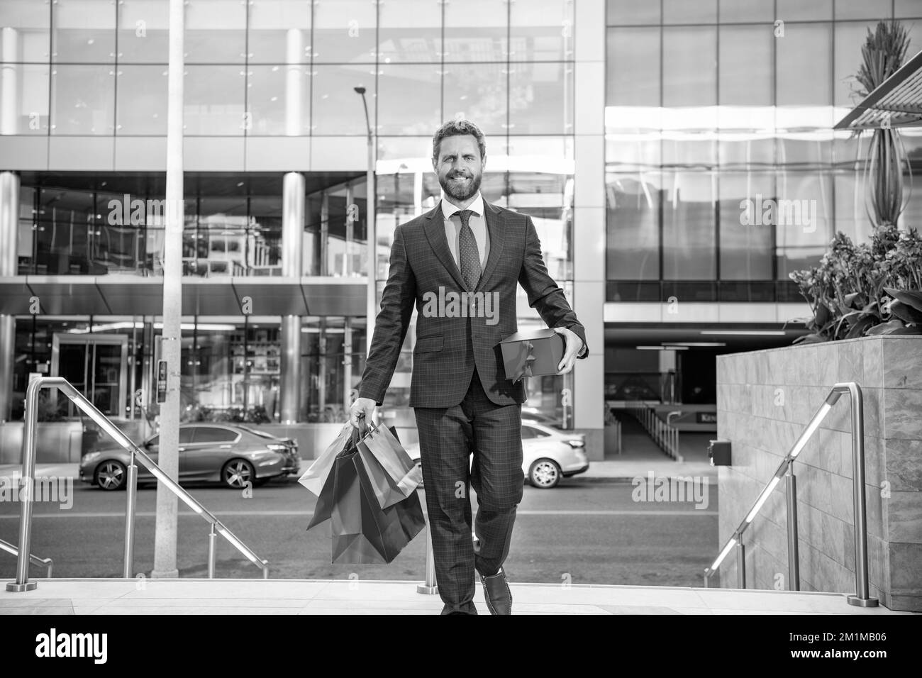 Glücklicher Unternehmer in Geschäftsanzug mit Einkaufstaschen Geschenke gehen vor dem Büro, Herrentag Stockfoto