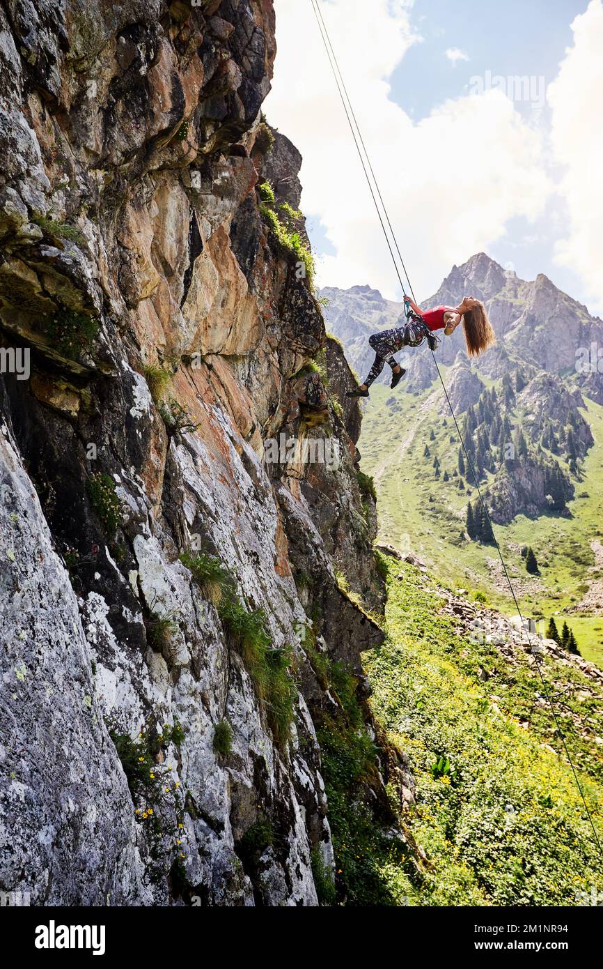 Fit, starke Kletterfrau, die in der Luft in der Nähe von hohen vertikalen Felsen mit Seilen in den Bergen Kasachstans fliegt Stockfoto