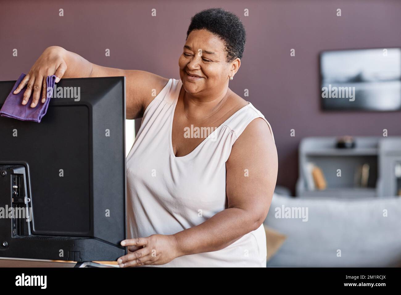 Porträt einer schwarzen Seniorin, die Fernsehen und gleichzeitig das gemütliche Zuhause putzt Stockfoto