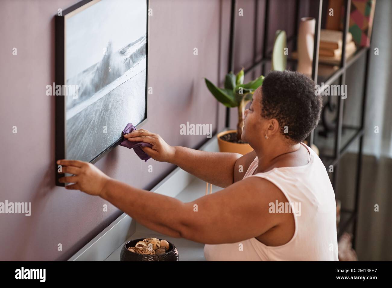 Seitliches Porträt einer schwarzen Seniorin, die Wandbilder abstaubt, während sie ein modernes Apartment putzt Stockfoto
