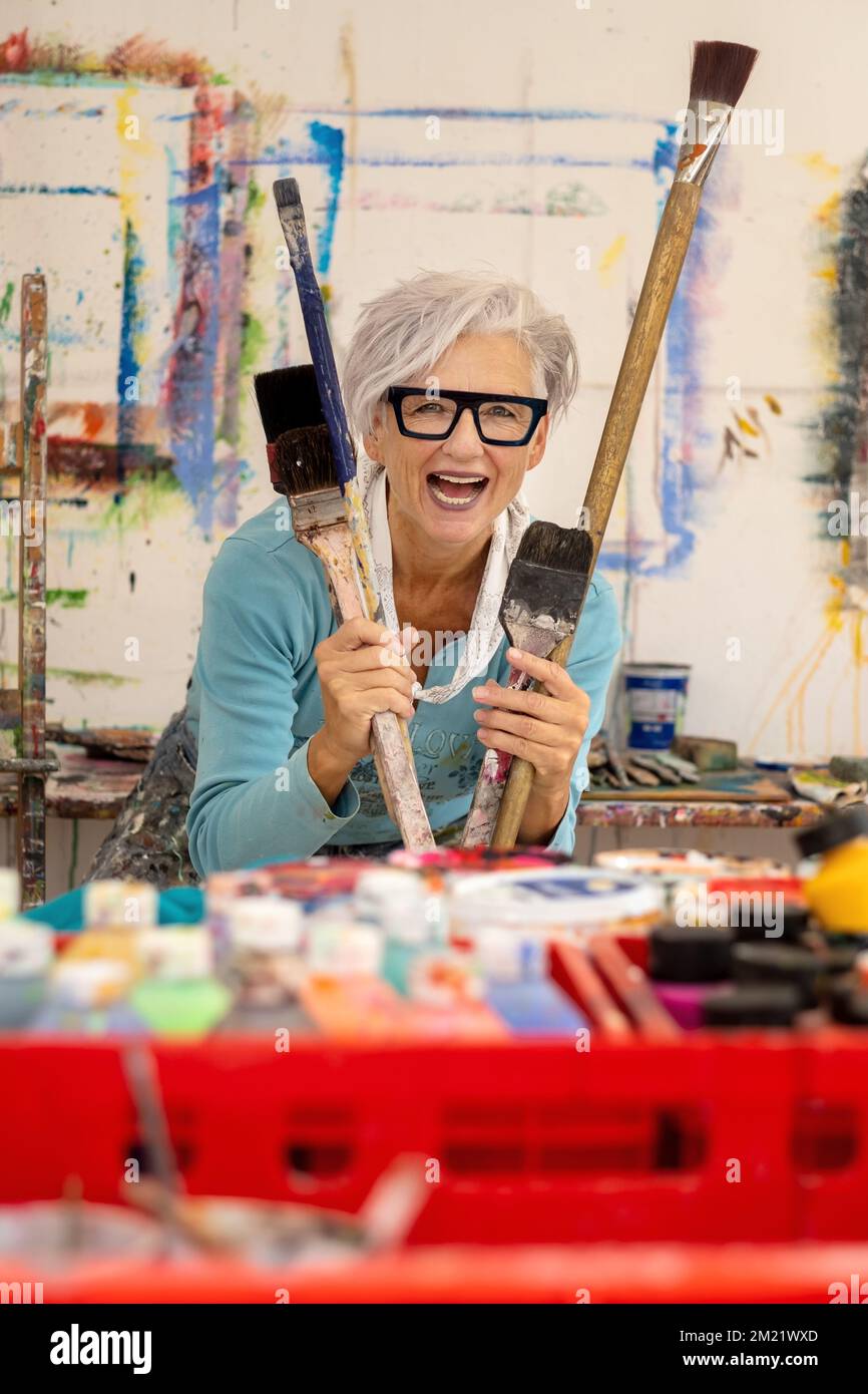 Ältere grauhaarige, reife, fröhliche, lachende Künstlerin mit Brille und großen Pinseln, in ihrem Atelier, Kopierraum Stockfoto
