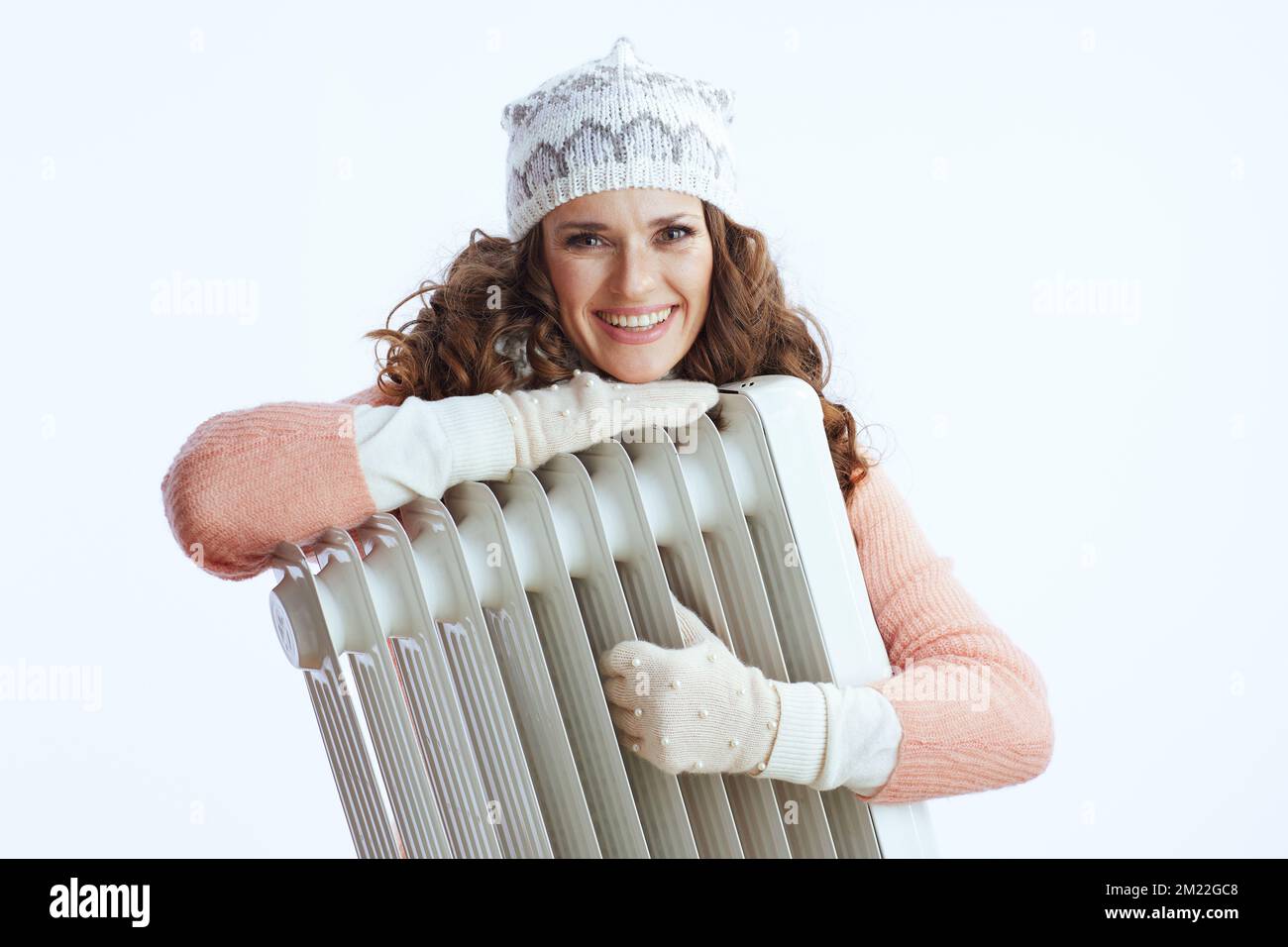 Hallo Winter. Lächelnde moderne Frau in Pullover, Fäustlingen, Hut und Schal vor weißem Hintergrund mit Heizung. Stockfoto