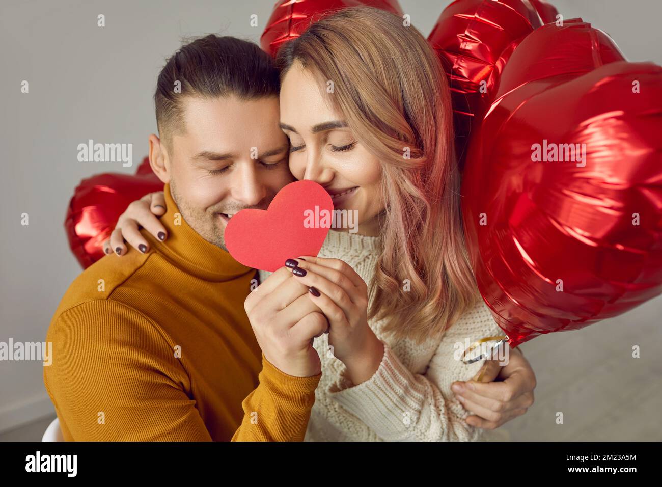 Portrait eines niedlichen, verliebten Paares, das mit geschlossenen Augen ein rotes Papierherz zusammenhält. Stockfoto