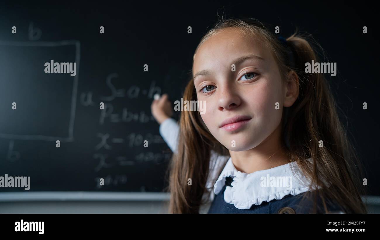 Porträt eines kaukasischen Mädchens im Klassenzimmer. Das Schulmädchen schreibt die Formel mit Kreide auf die Tafel und schaut in die Kamera. Stockfoto