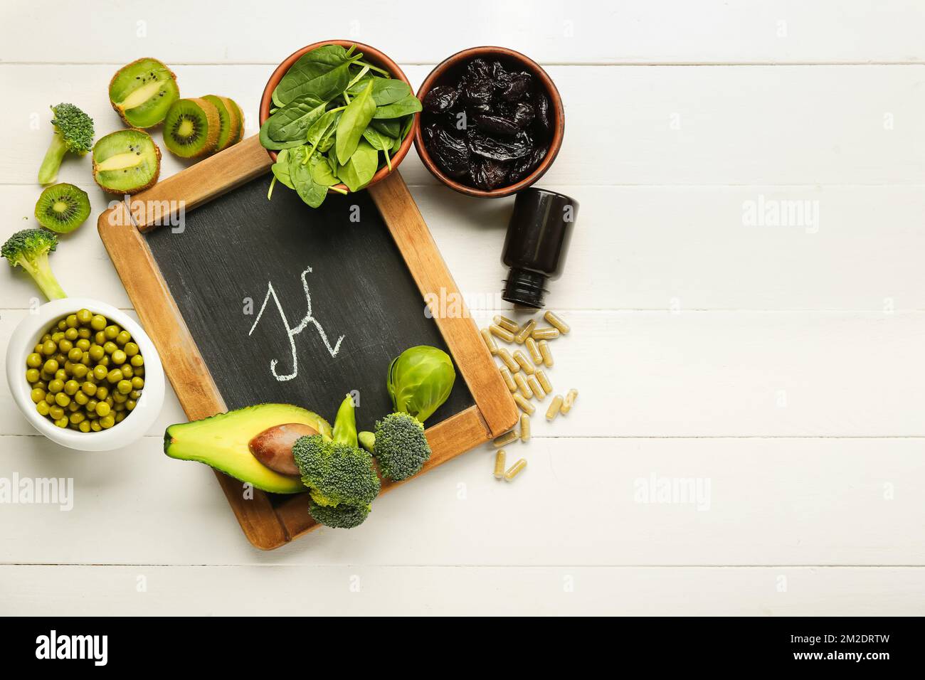 Tafel mit dem Buchstaben K, gesunde Produkte und eine Flasche Pillen auf weißem Holzhintergrund Stockfoto