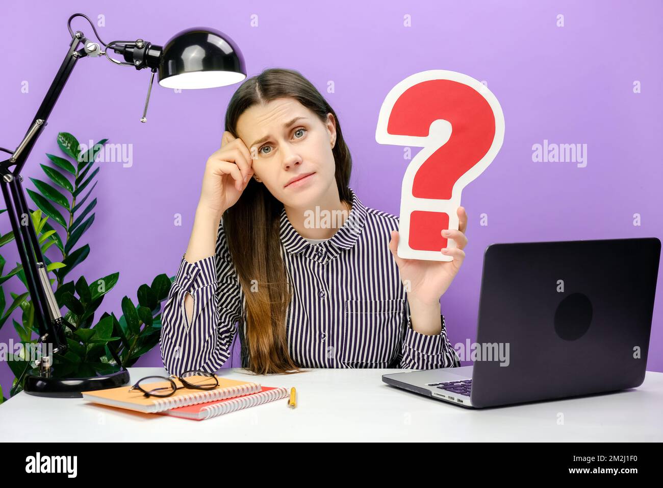 Eine pensive, traurige junge Mitarbeiterin im Alter von 20s Jahren, die am weißen Büroschreibtisch-pc-Laptop sitzt und ein rotes Fragezeichen hält und isoliert über violett posiert Stockfoto