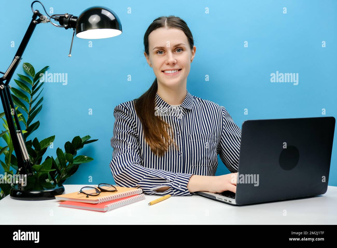 Portrait einer glücklichen, wunderschönen, erfolgreichen Sekretärin, junge Frau 20s, die am weißen Büroschreibtisch mit pc-Laptop-Computer sitzt und isoliert über Paste posiert Stockfoto