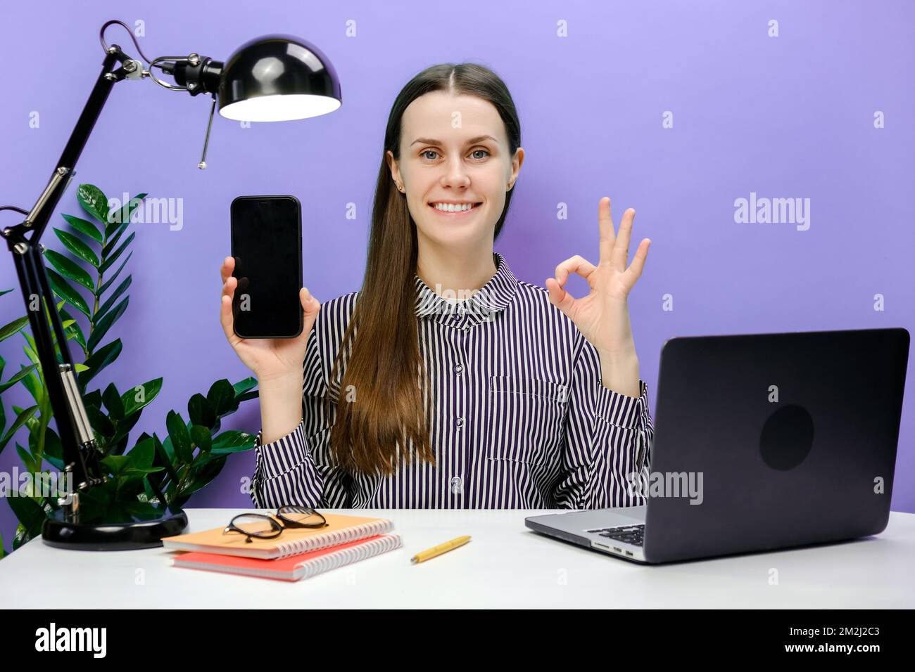 Erfolgreiche Mitarbeiterin, junge Frau in legerem Hemd, die am Büroschreibtisch sitzt, mit einem Laptop, der ein Mobiltelefon benutzt, eine gute Geste zeigt, isoliert über dem Schreibtisch posiert Stockfoto
