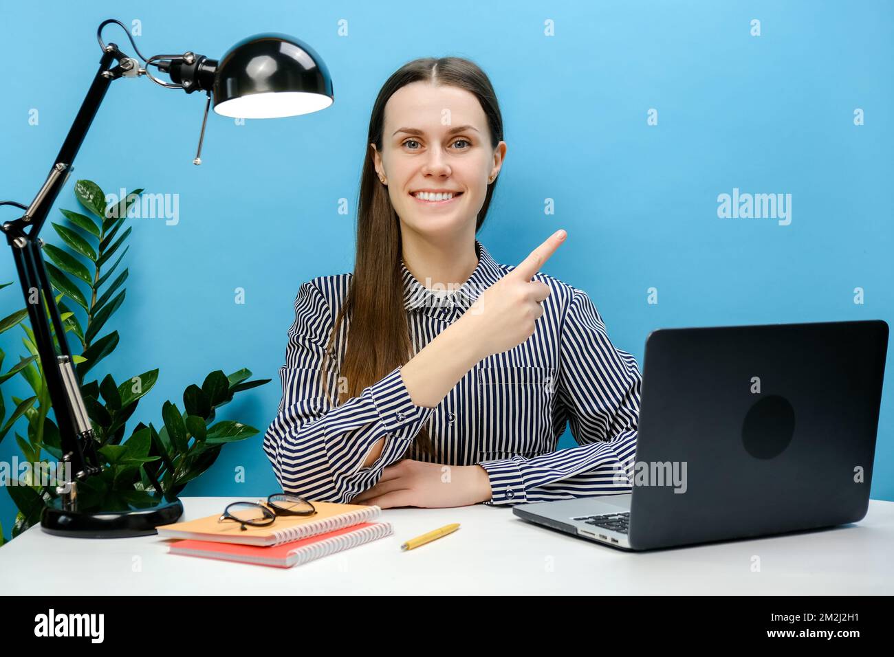Glückliche erfolgreiche Mitarbeiterin Geschäftsfrau mit 20s in einem legeren Hemd sitzt am weißen Büroschreibtisch mit Laptop Zeigefinger beiseite auf Kopierfeld, posiert Stockfoto