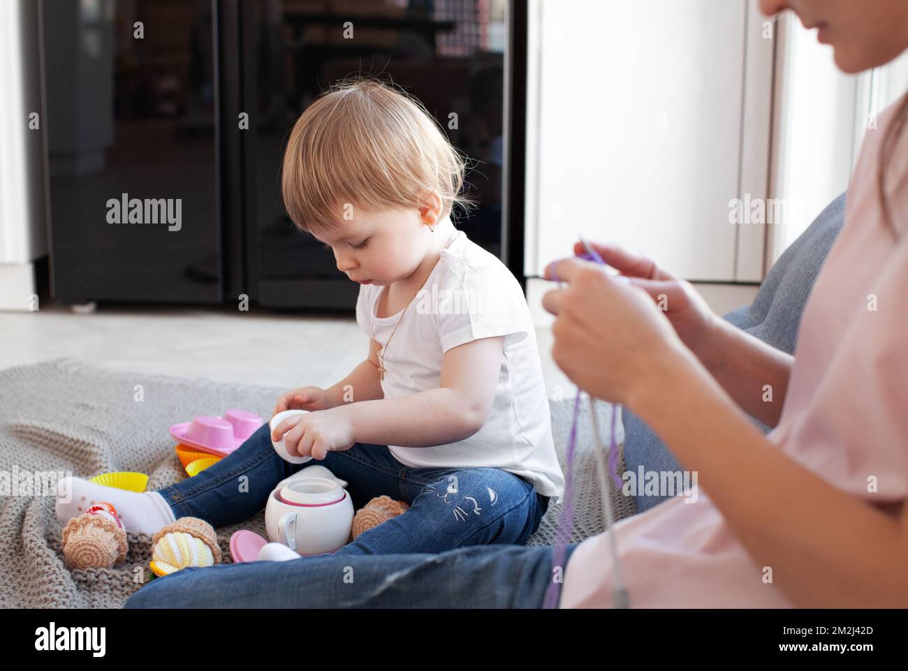Kleine Tochter spielt, während ihre Mutter strickt, Arbeit oder Hobby macht. Familie, ehrliches Familienleben mit Baby. Authentischer Lifestyle Stockfoto