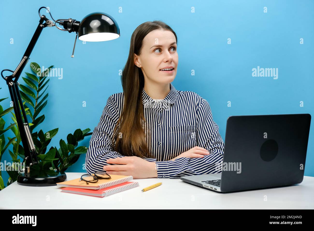 Porträt einer geistesbewussten, nachdenklichen Geschäftsfrau mit 20s-cm-Hemd, die auf einem weißen Büroschreibtisch mit pc-Laptop sitzt und isoliert über einem einfarbigen blauen Hintergrund posiert Stockfoto