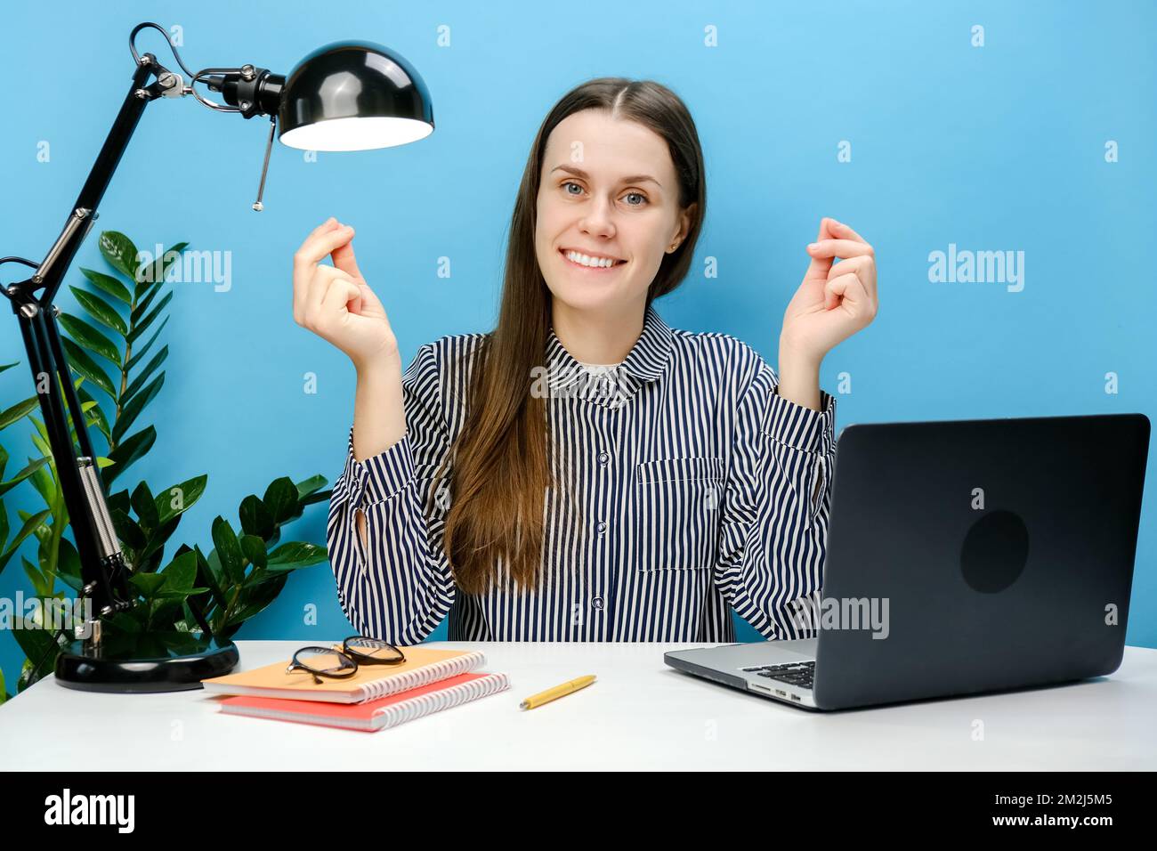 Lächelnde, erfolgreiche Mitarbeiterin in einem legeren Hemd, die am weißen Büroschreibtisch sitzt und ihre Finger am Laptop reibt, zeigt, dass sie nach Geld fragt, isoliert Stockfoto