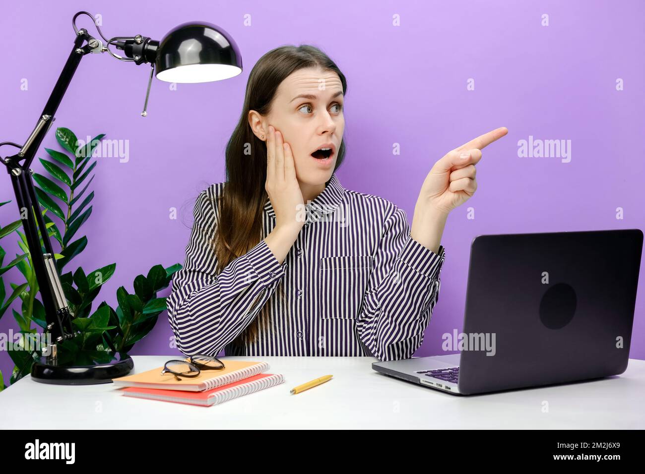 Schockierte, erfolgreiche Mitarbeiterin Geschäftsfrau 20s sitzt am weißen Büroschreibtisch mit Laptop Zeigefinger beiseite auf Arbeitsbereich halten Kopf, posieren Stockfoto