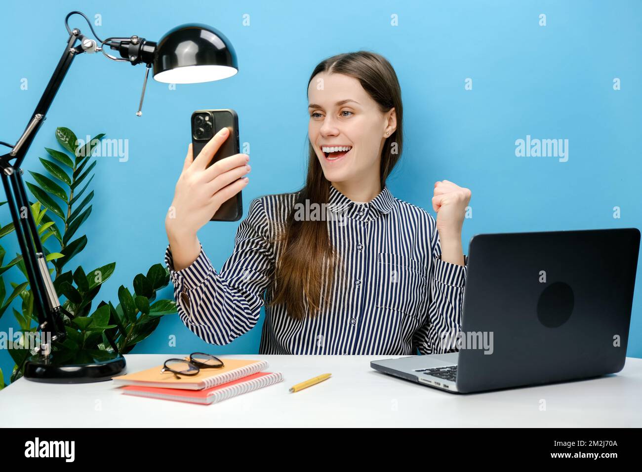 Portrait einer erfolgreichen Mitarbeiterin, einer jungen Geschäftsfrau im Hemd, die am weißen Büroschreibtisch sitzt und ein Laptop mit Mobiltelefon hält, die Sieger-Gestenklausel Stockfoto