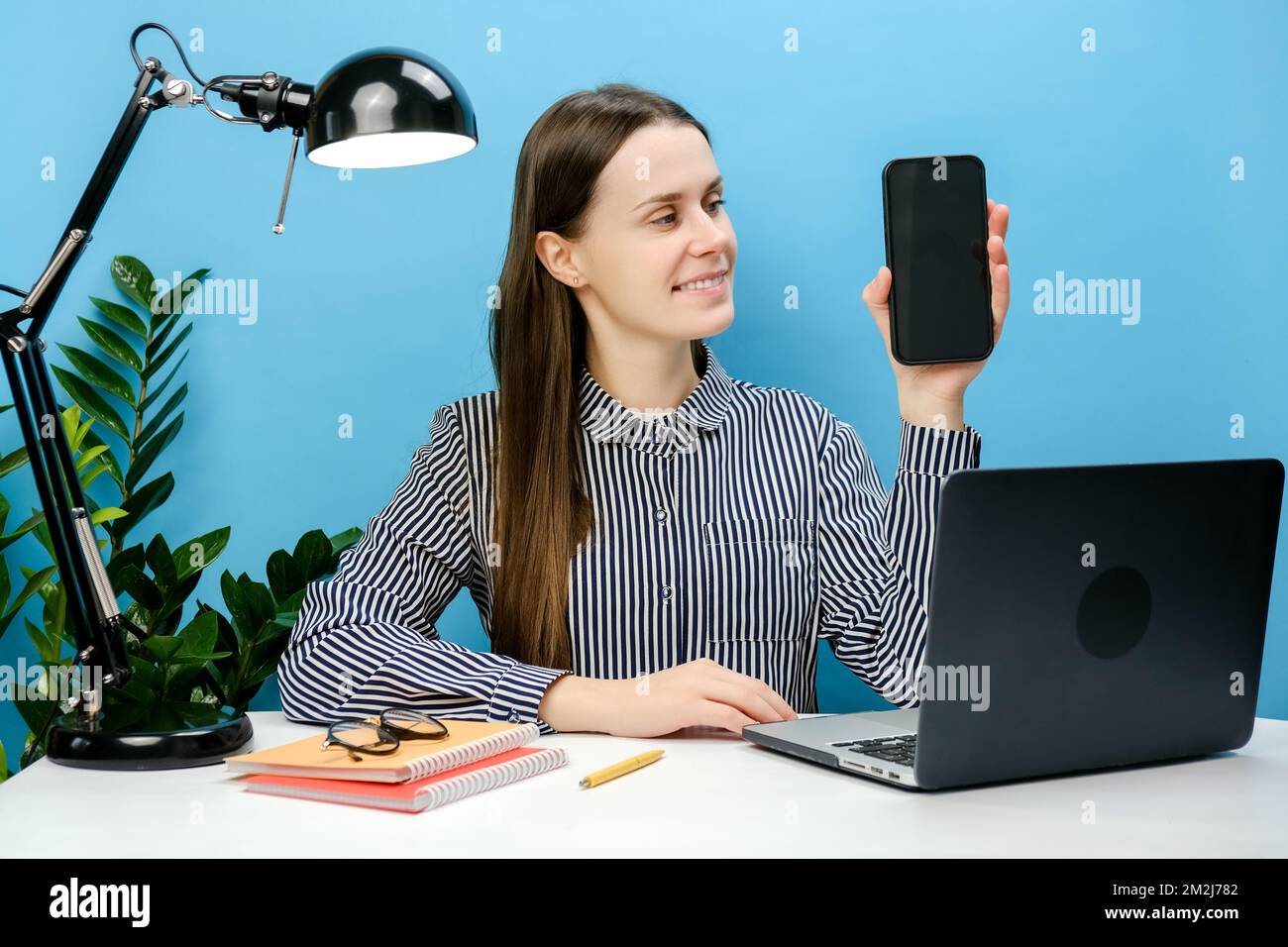 Glückliche, erfolgreiche Geschäftsfrau im Hemd, die am weißen Büroschreibtisch sitzt und einen Laptop mit einem Mobiltelefon mit leerem Bildschirm im Arbeitsbereich hat, Stockfoto