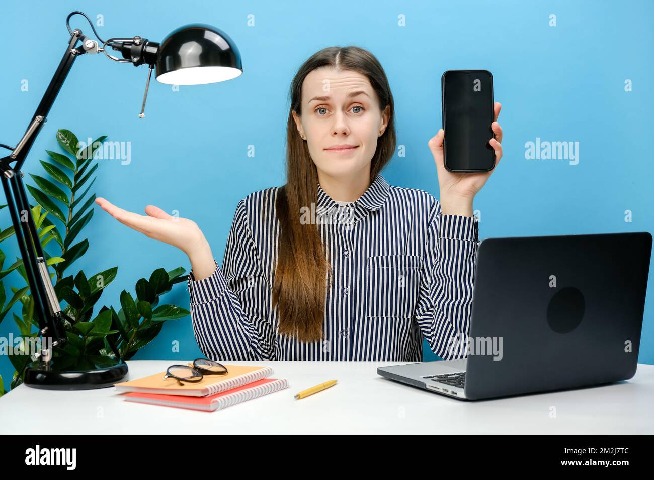 Verwirrte, traurige Geschäftsfrau, die am weißen Büroschreibtisch sitzt und ein Notebook mit einem Mobiltelefon hält, einen leeren Bildschirm im Arbeitsbereich hat die Hände verteilt Stockfoto