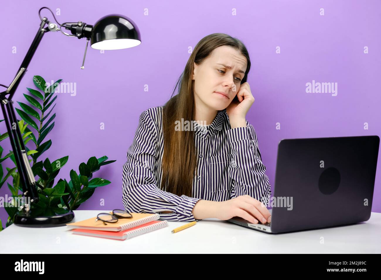 Porträt einer müden, frustrierten Sekretärin, einer jungen Frau in einem lässigen Hemd, die am weißen Büroschreibtisch sitzt und ein Laptop-Internet-Requisit verwendet Stockfoto
