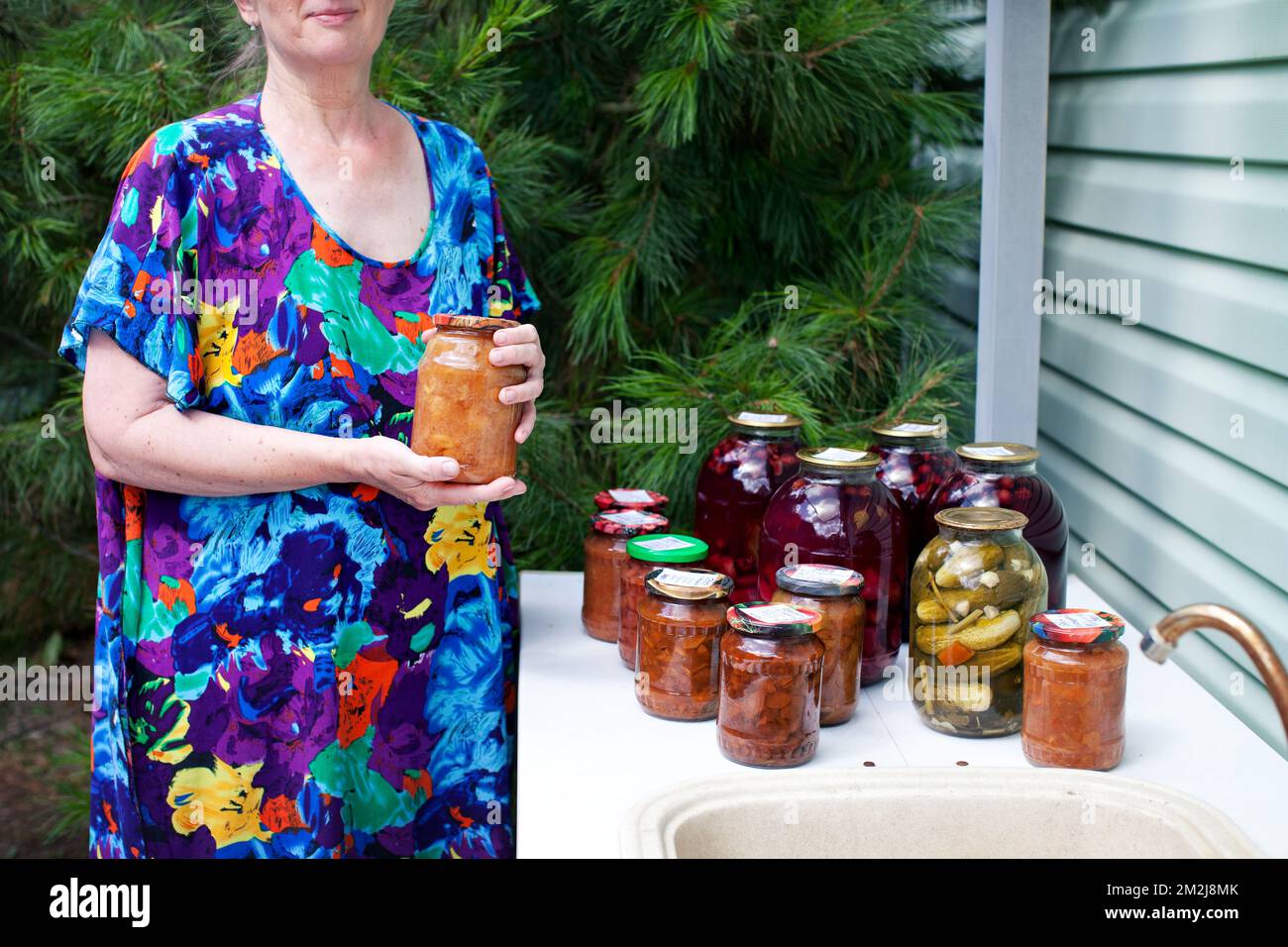 Eine alte Frau hält ein Glas hausgemachter Marmelade in den Händen. Das ältere Modell in blauem Kleid auf dem Hintergrund handwerklich hergestellter Konservendosen mit eingelegten Gurken Stockfoto