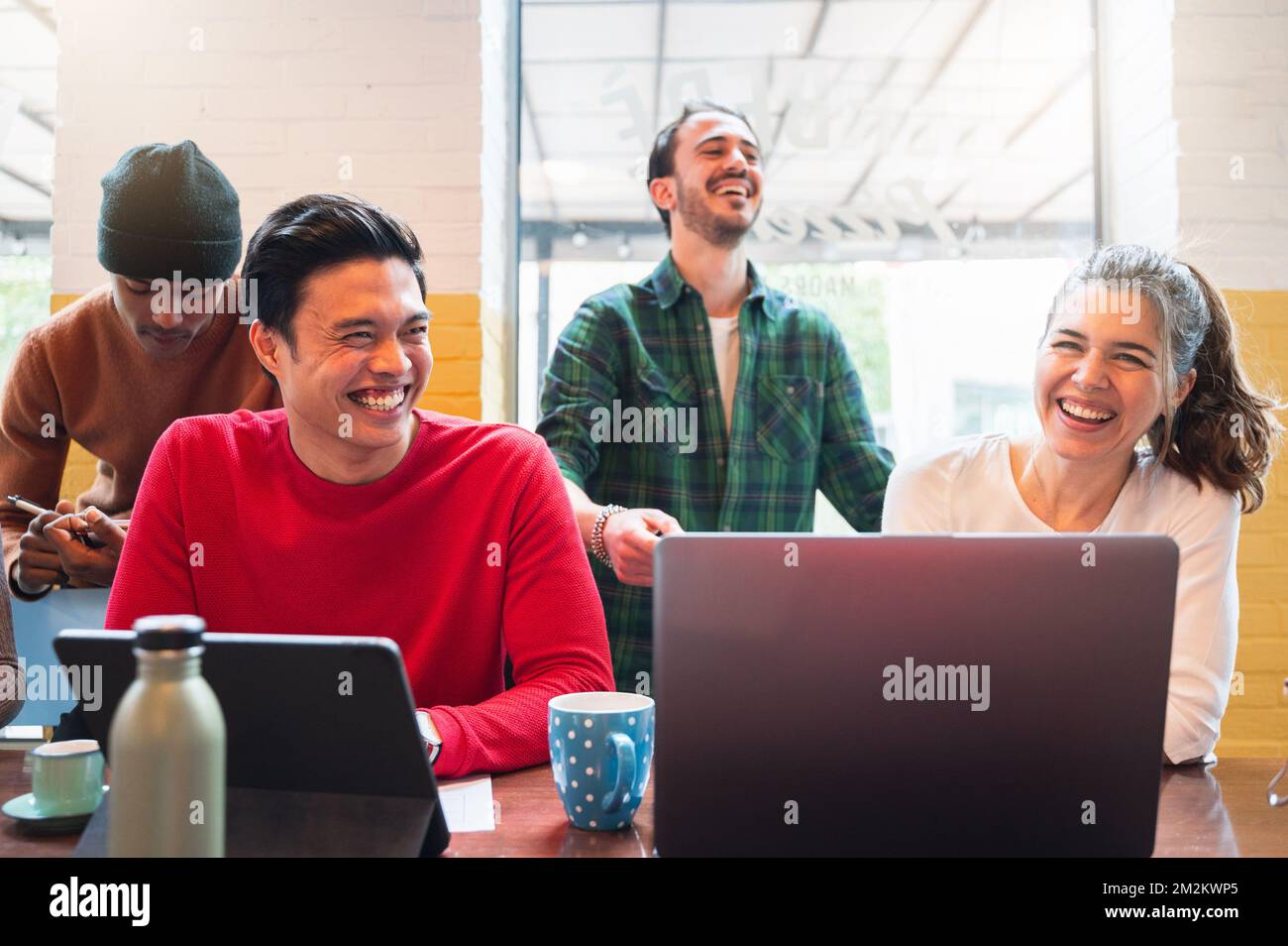 Gruppe von vier multiethnischen Jugendlichen, die am Tisch sitzen und Computer benutzen. Männer, die beim Arbeiten oder Lernen lachen. Zusammenarbeit, Einheit und Zusammengehörigkeit Stockfoto
