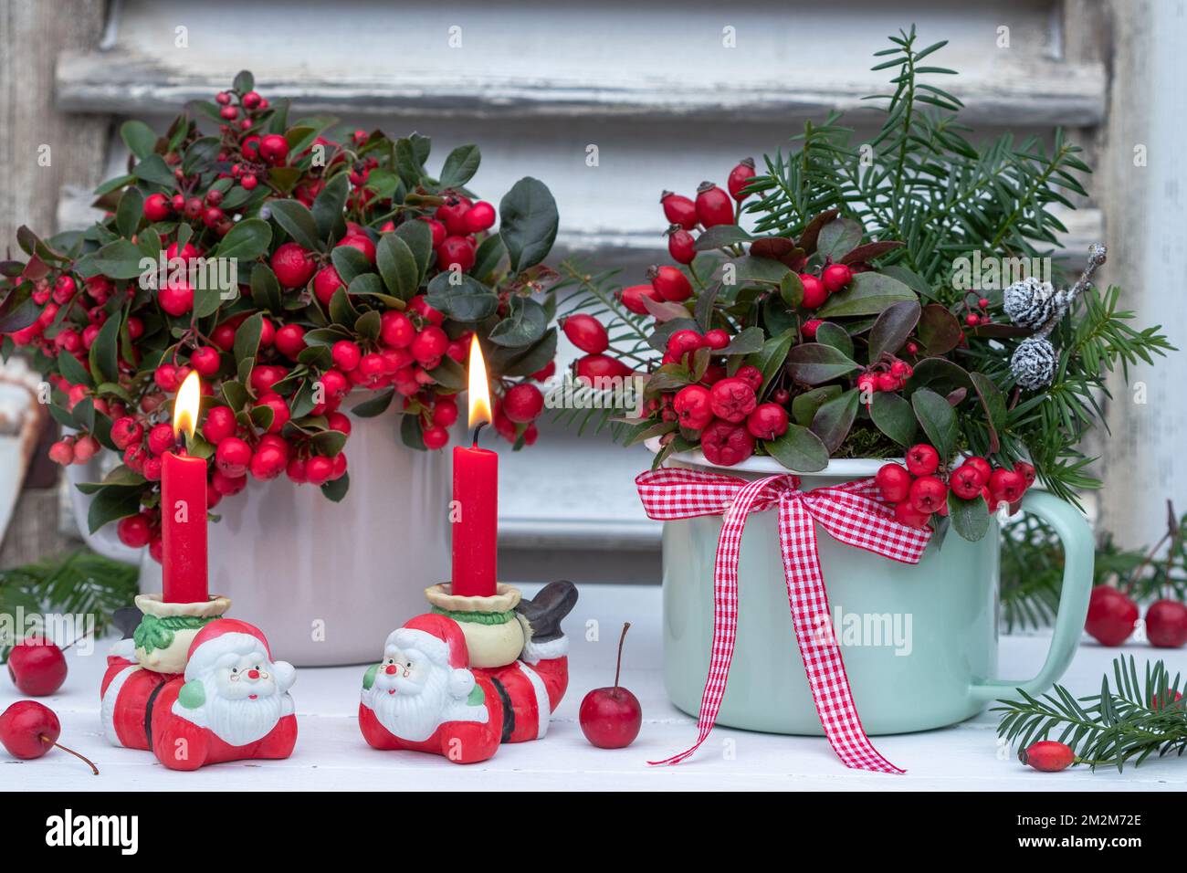 weihnachtsdekoration mit roten Kerzen, Weihnachtsmann und amerikanischem Wintergrün in emaillierten Töpfen Stockfoto