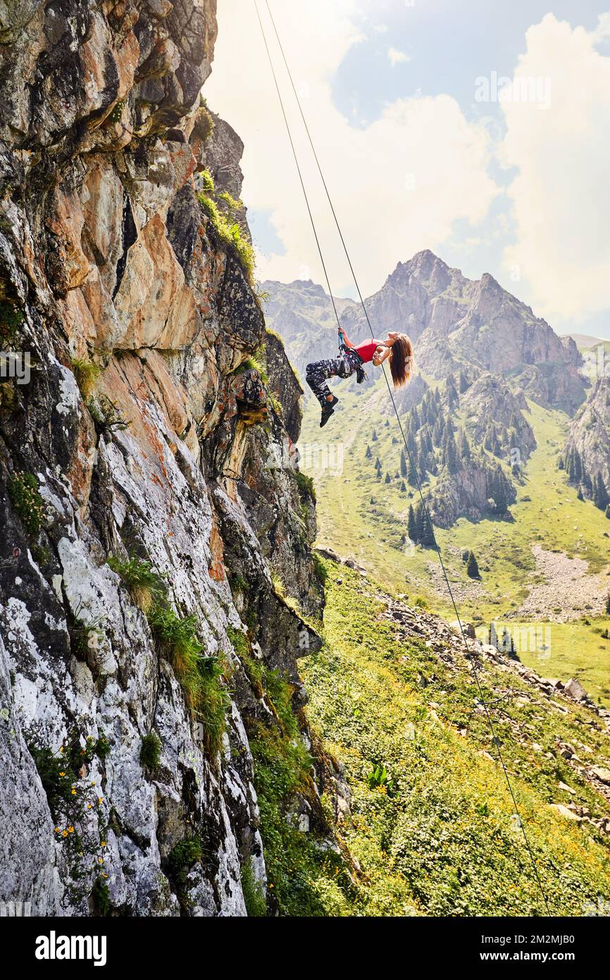 Stecke eine starke Klettererin in ein rotes Hemd mit langen Haaren, die in der Luft in der Nähe von hohen vertikalen Felsen mit einem Seil in den Bergen Kasachstans fliegen Stockfoto