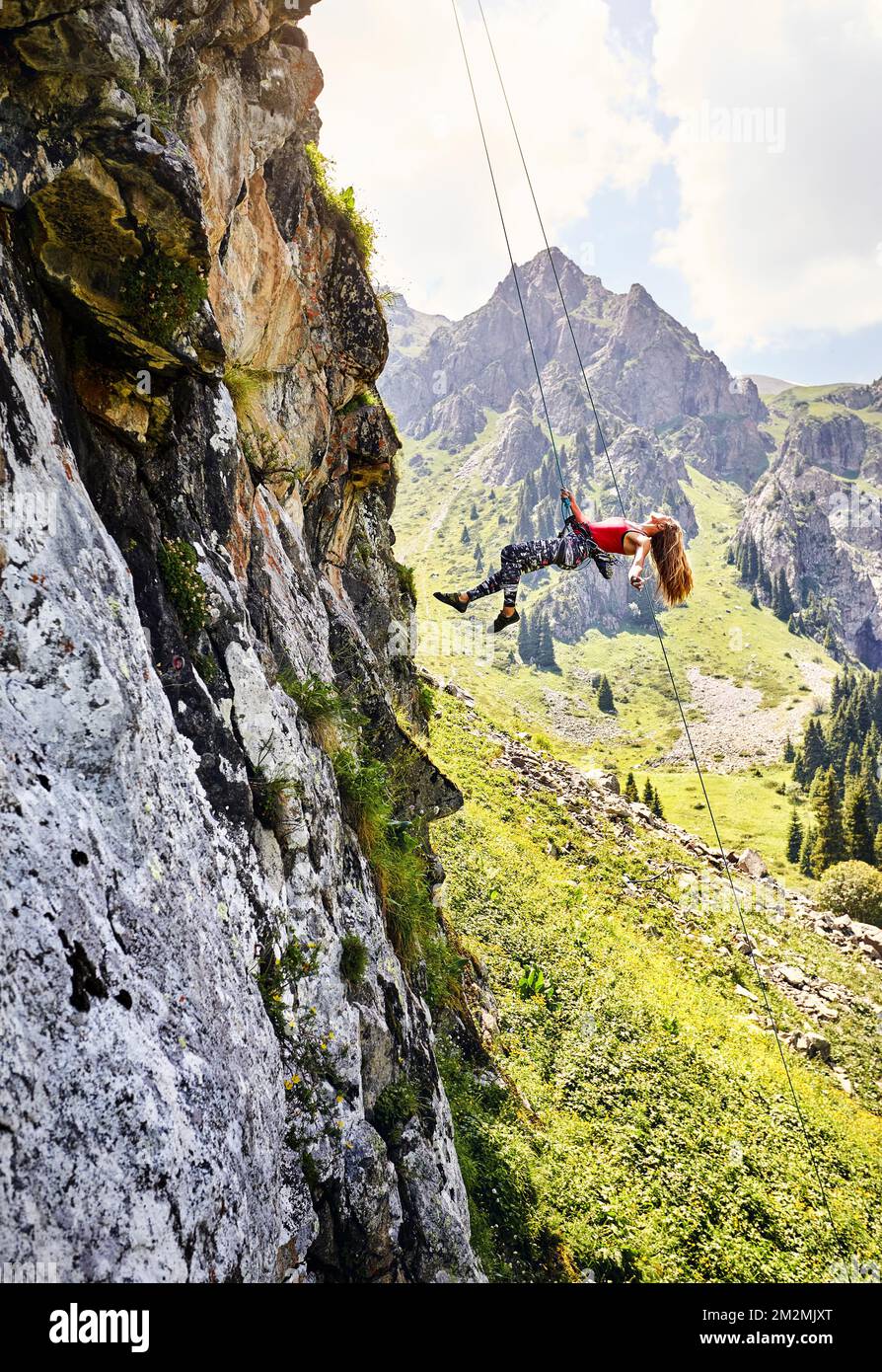 Stecke eine starke Klettererin in ein rotes Hemd mit langen Haaren, die in der Luft in der Nähe von hohen vertikalen Felsen mit einem Seil in den Bergen Kasachstans fliegen Stockfoto