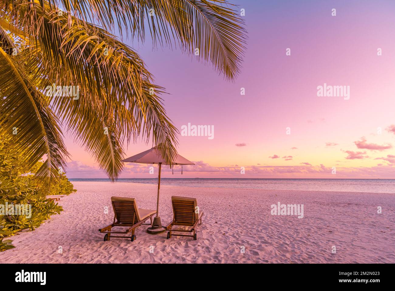 Panoramablick auf den tropischen Sonnenuntergang, zwei Sonnenliegen, Sonnenschirme unter Palmen. Weißer Sand, Blick auf den Meershorizont, farbenfroher Sonnenuntergang am Himmel Stockfoto