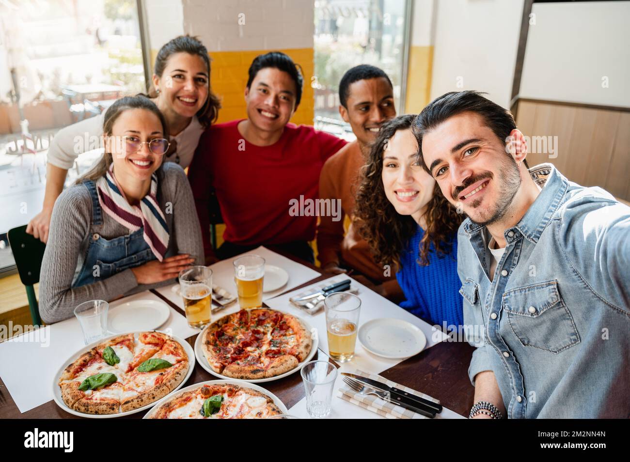 Eine multiethnische Gruppe von sechs Freunden macht ein Selfie-Foto in einem Restaurant am Tisch, während sie Pizza essen. Menschen mit fröhlichem, fröhlichem Gesichtsausdruck. Stockfoto