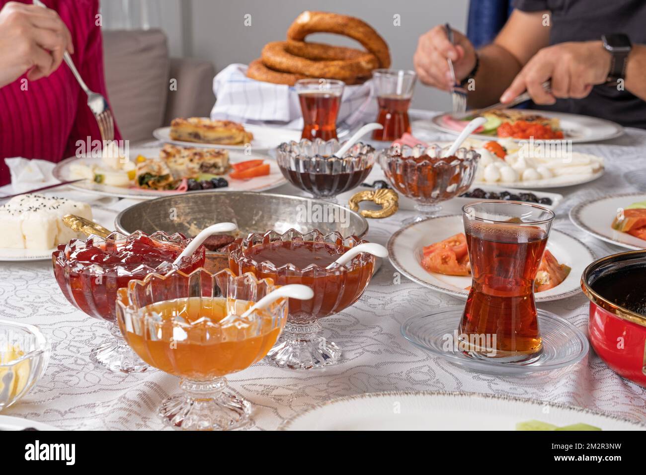 Zugeschnittenes Foto eines reichen Familienfrühstücks. Ich trinke heißen, frischen türkischen Tee im Glas. Männliche und weibliche Hand mit Löffel und Gabel im Hintergrund. Stockfoto