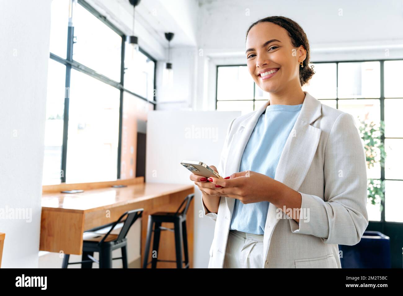 Foto einer positiven hispanischen oder brasilianischen Frau, Unternehmerin, die in einem modernen Büro steht, ihr Mobiltelefon nutzt, online mit einem Kollegen oder Kunden kommuniziert, in die Kamera schaut und freundlich lächelt Stockfoto