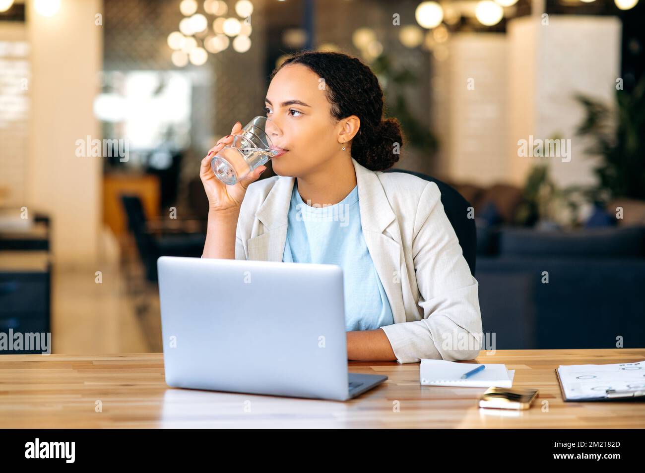 Gesunde brasilianische oder hispanische Geschäftsfrau, sitzt an einem Arbeitsplatz im Büro, trinkt ein Glas frisches sauberes Wasser, hält das Gleichgewicht des täglichen Wassers. Gesundheitsfürsorge, tägliche Wasseraufnahme im Körper Stockfoto