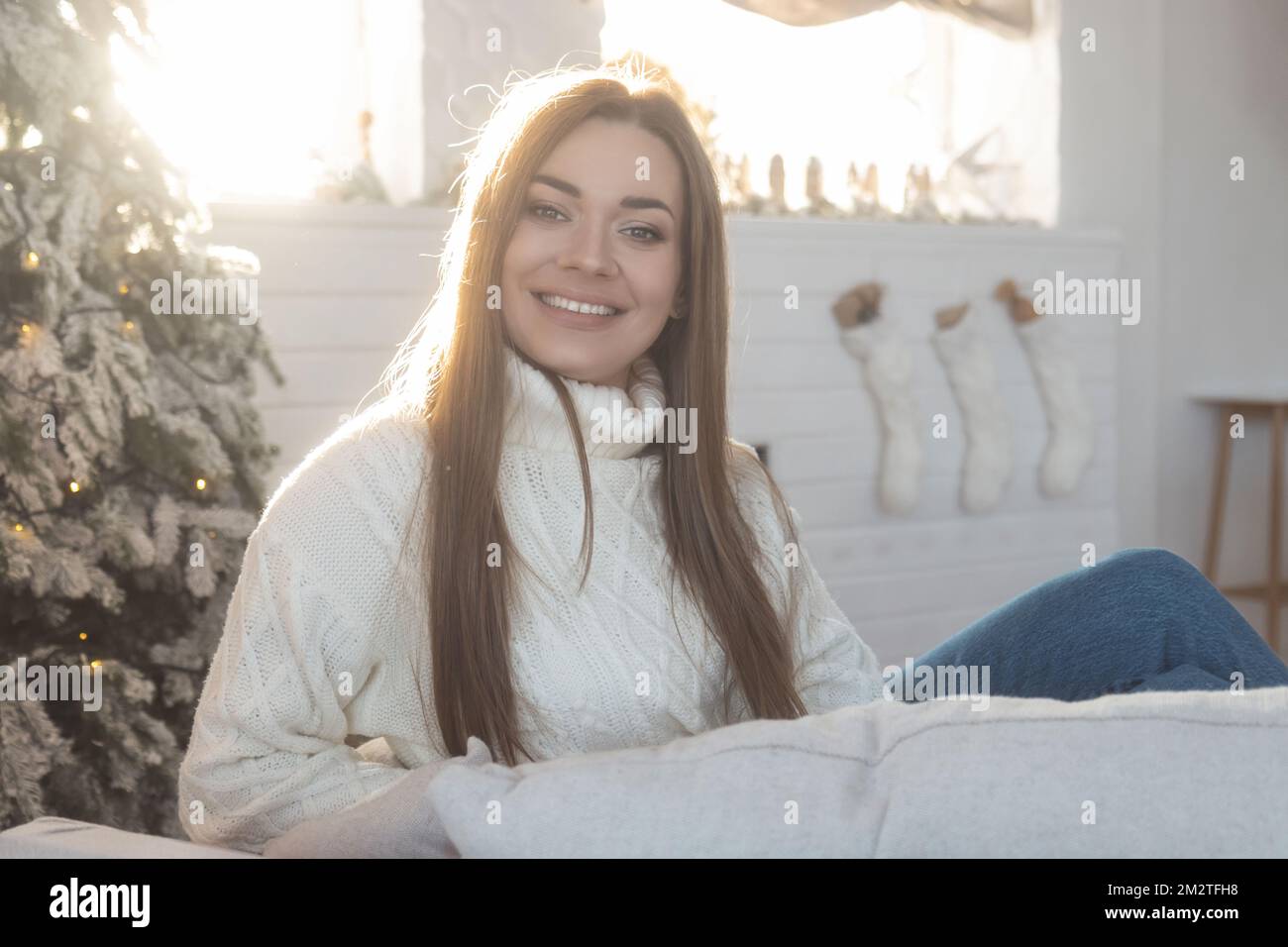 Ein hübsches Mädchen in einem weißen Pullover vor dem Hintergrund der Neujahrsdekoration sitzt auf einem Stuhl Stockfoto