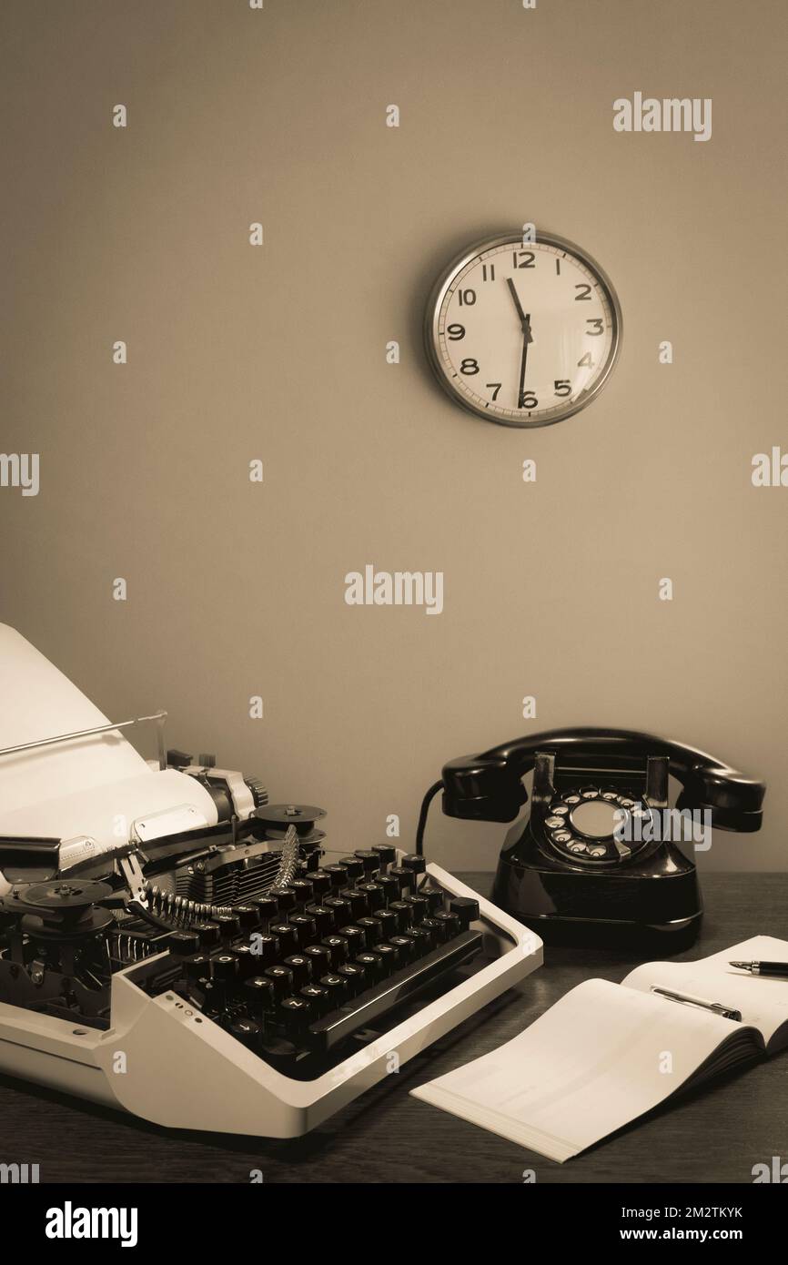 Eine Retro-Schreibmaschine und ein altes Telefon liegen auf dem Tisch, zusammen mit einem Scheckbuch und einem Tintenstift. Uhr an der Wand. Schwarz auf Weiß Stockfoto