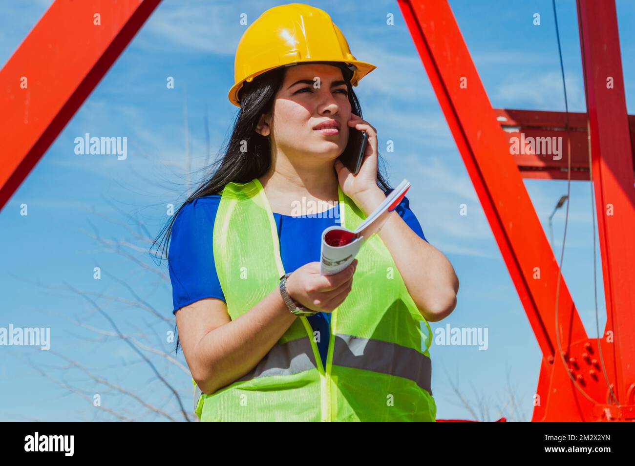 Junge, weiße Latina-Ingenieurin im Freien, trägt einen gelben Schutzhelm und eine Sicherheitsweste, verwirrt bei einem Anruf, der mit ihrem Boss spricht, hält Workboo Stockfoto