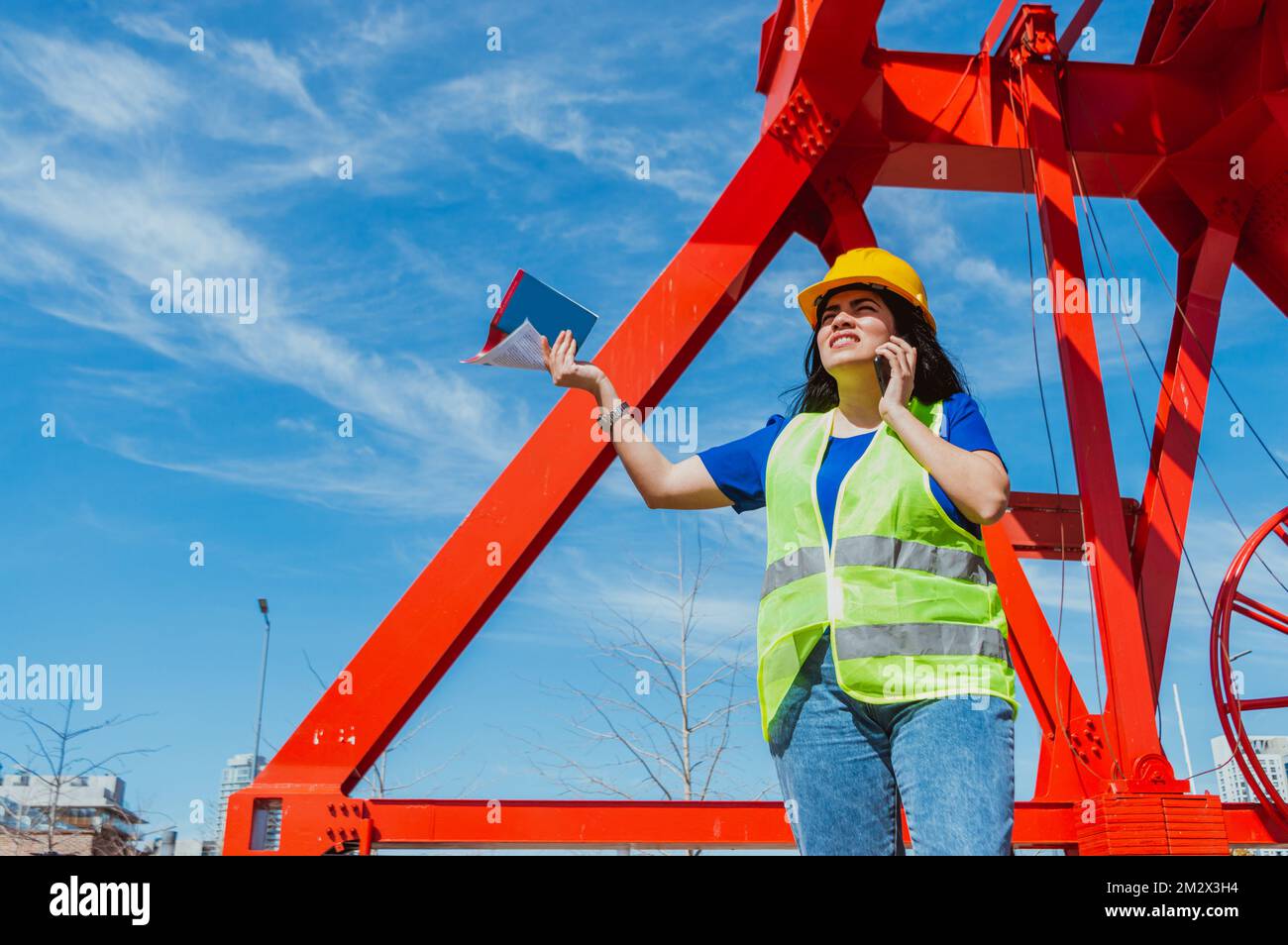 Junge lateinweiße Ingenieurin, trägt Schutzhelm und Sicherheitsweste, steht auf einer Baustelle, streitet sich am Telefon und hält Workbo Stockfoto