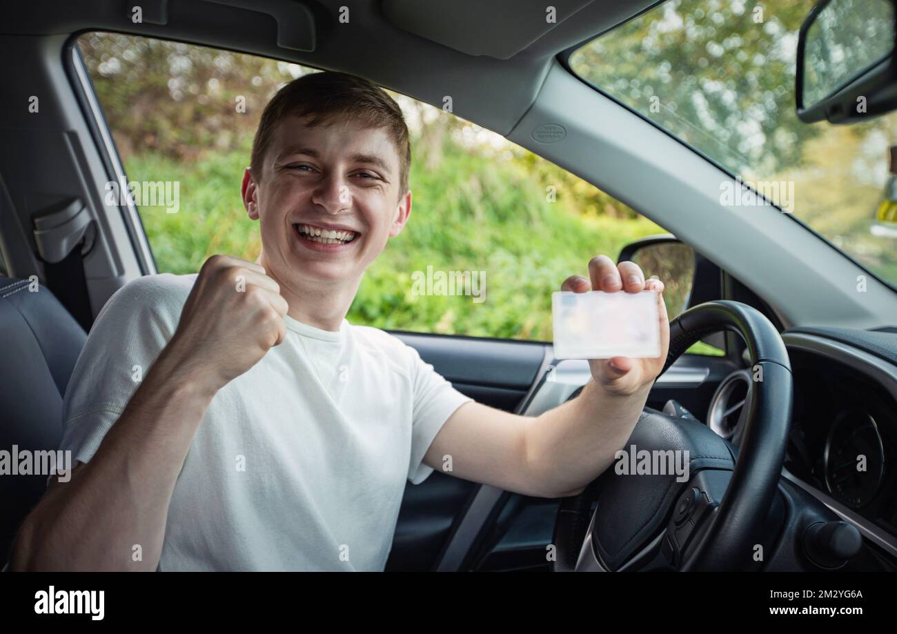 Glücklicher und stolzer Kerl, der seinen Führerschein aus dem Autofenster zeigt, hält die Faust fest als Sieger, der den Sieg feiert. Bestehen des Tests und Fahren Stockfoto