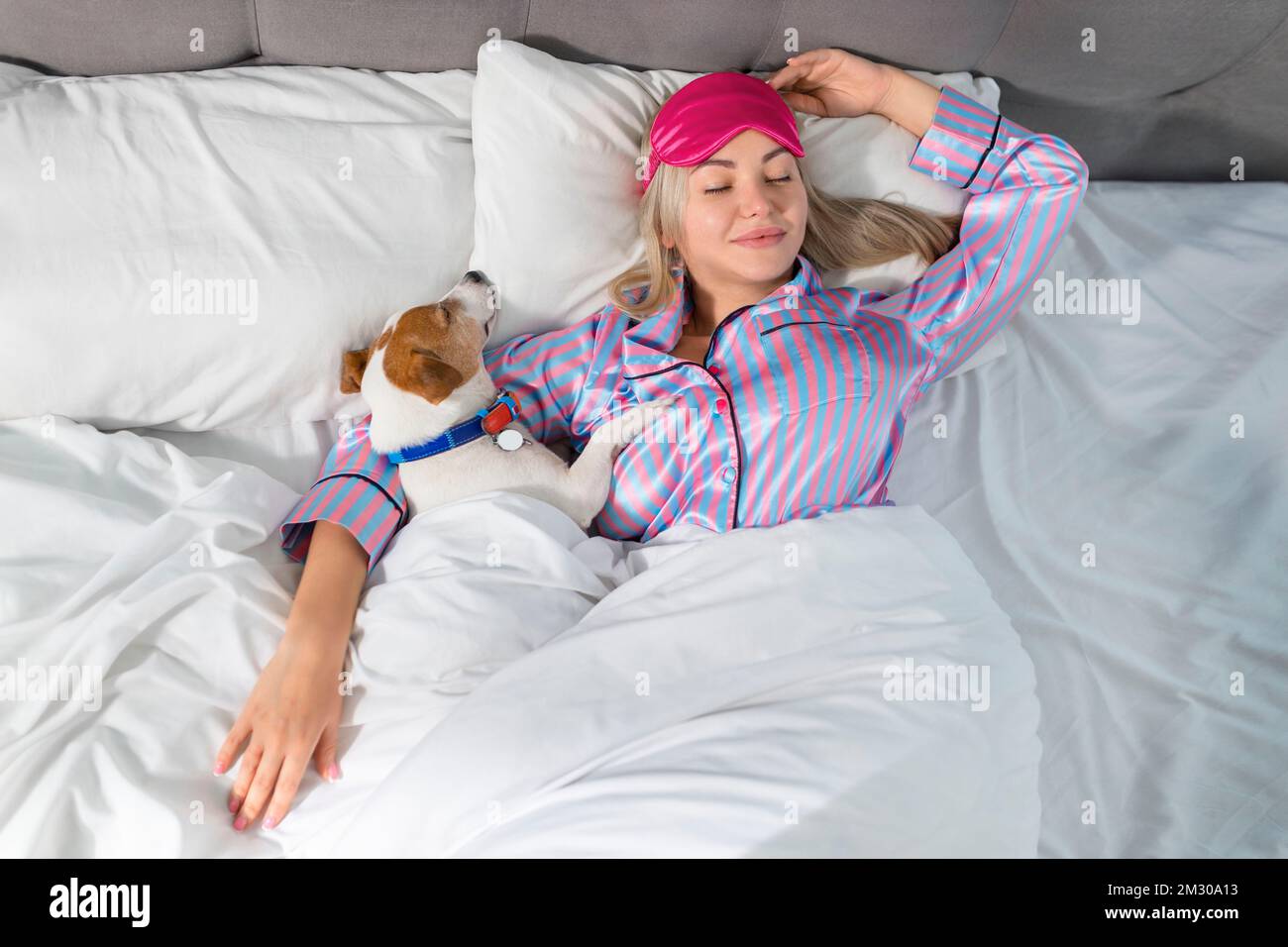 Eine attraktive junge Frau im Pyjama hält einen Hund, während sie auf einem Bett liegt Stockfoto