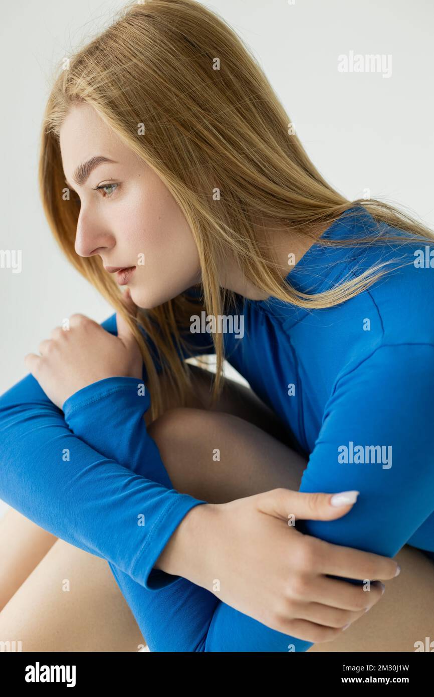 Seitenansicht einer Frau in hellblauem Rollkragenpullover, die sich an den Knien anschließt, während sie isoliert auf einem grauen Stockbild sitzt Stockfoto