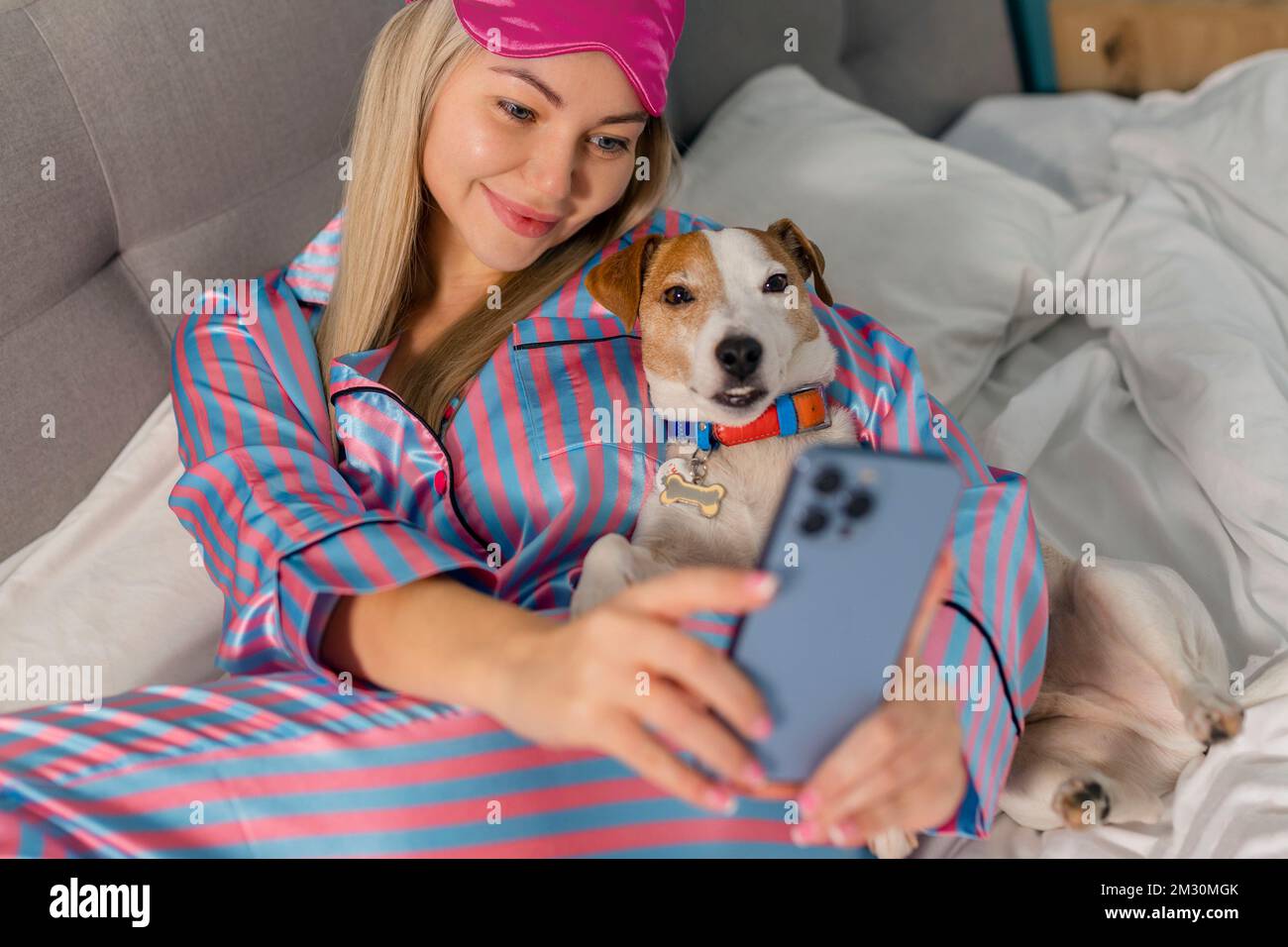 Porträt einer schönen, fröhlichen Frau im Pyjama, die den Hund umarmt und mit dem Handy ein lächelndes Selfie macht, während sie nach dem Schlafen oder Nickerchen auf dem Bett liegt Stockfoto