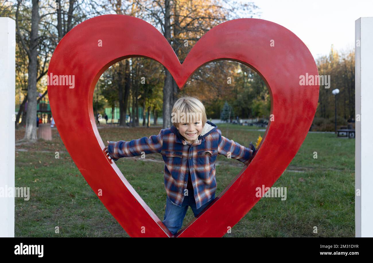 Ein süßer, lächelnder Junge, 5-6 Jahre alt, in einem karierten Hemd in einem großen roten Herz, sieht glücklich in der Kamera aus. Muttertag, valentinstag Stockfoto