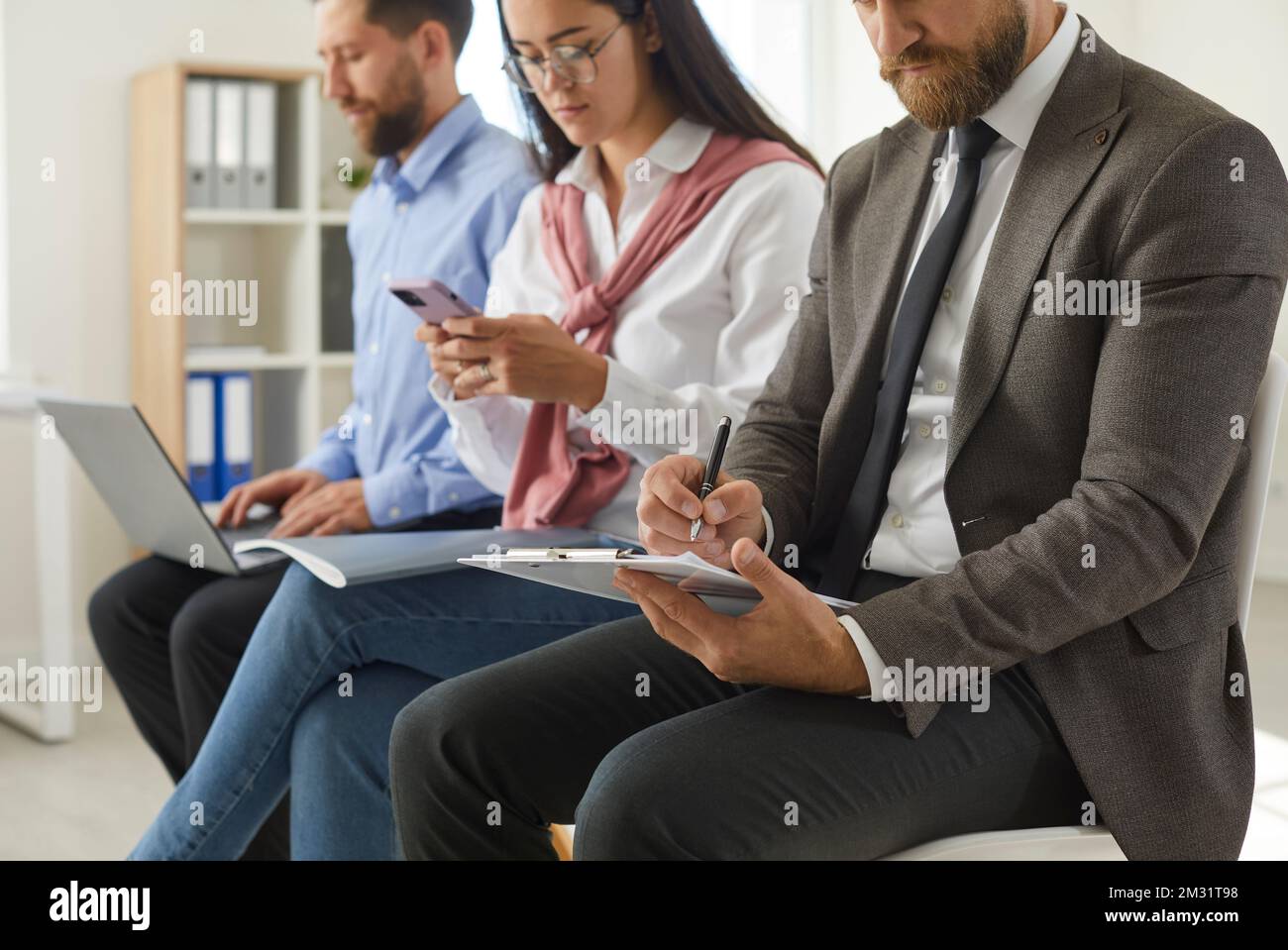 Geschäftsleute, die im Büro sitzen, auf Klemmbrettern schreiben und Laptops und Mobiltelefone verwenden Stockfoto
