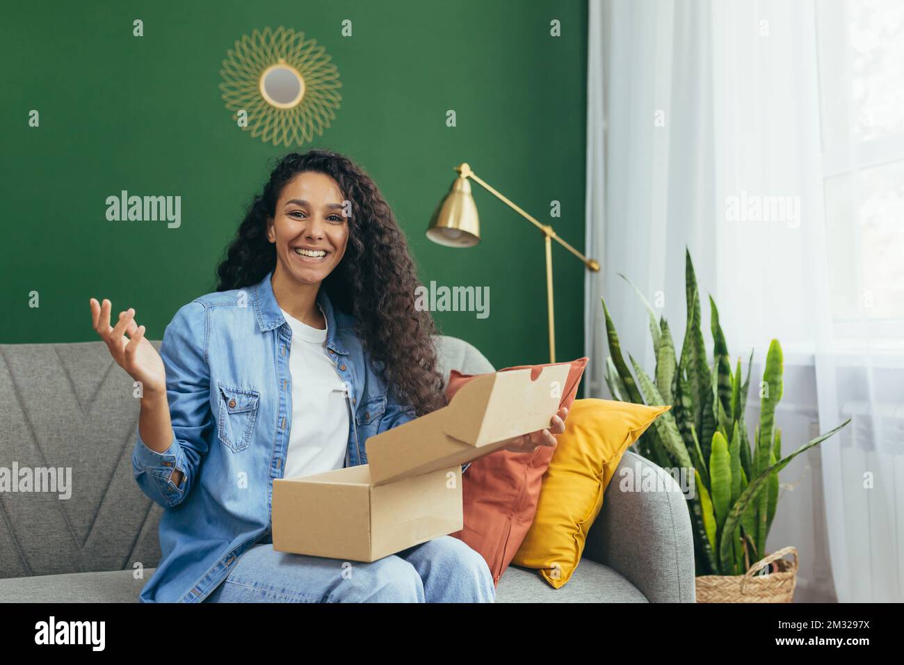 Glückliche Käuferin zu Hause zufrieden mit der Lieferung von Waren aus dem Online-Shop, hispanische Frau, die auf dem Sofa sitzt und mit Waren in der Hand auf eine Kamera schaut. Stockfoto