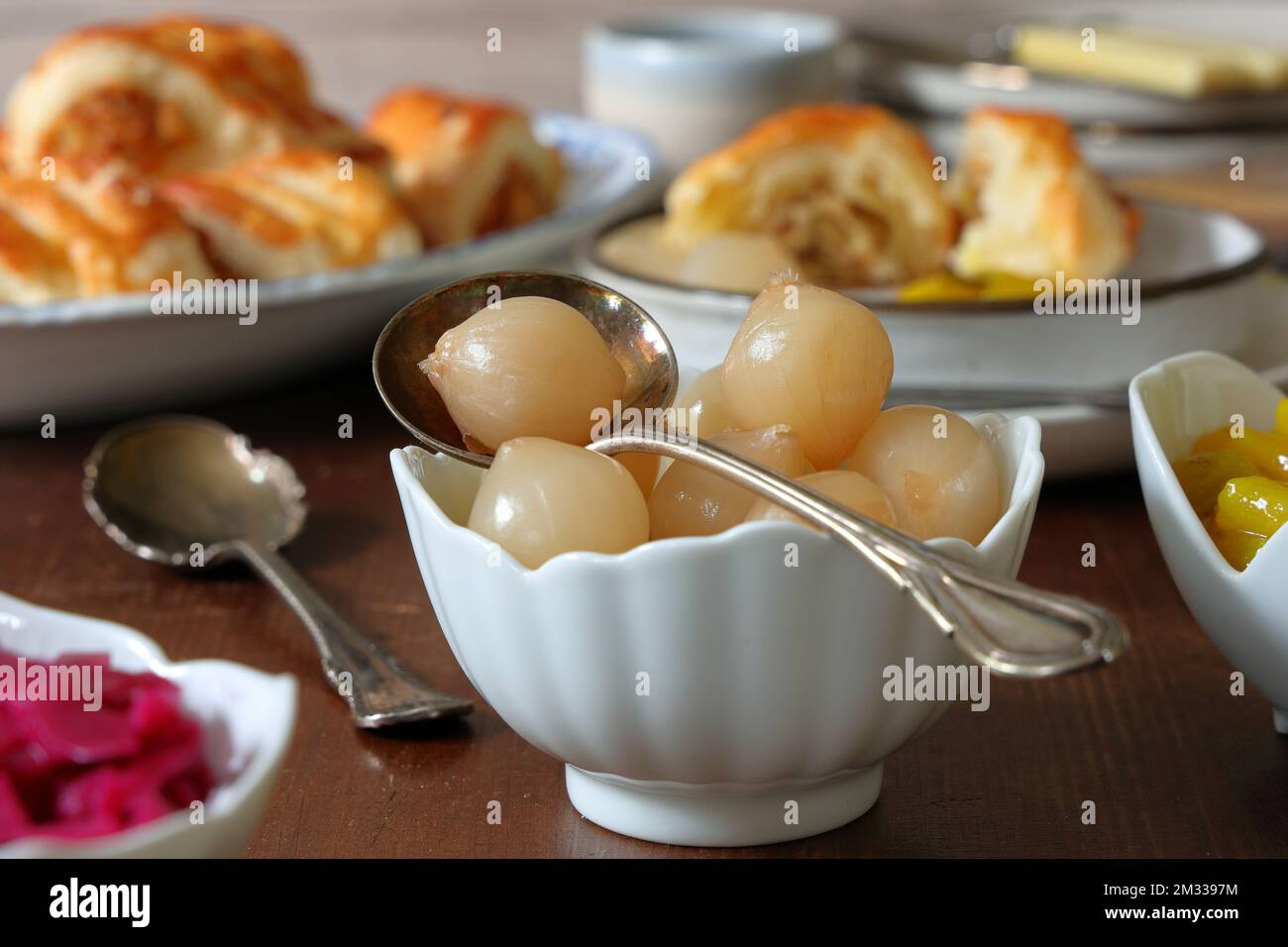Eine Schüssel eingelegter silberner Zwiebeln mit traditionellen dekorativen Löffeln auf einem dunklen Holztisch mit Schüsseln anderer Gurken und serviert mit herzhaftem Gebäck Stockfoto